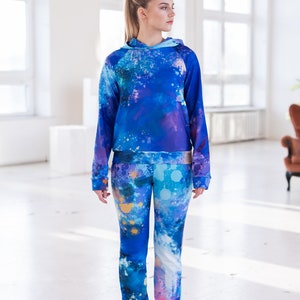 May include: A blue and purple hooded sweatshirt and matching pants with a paint splatter design. The model is wearing pink sneakers.