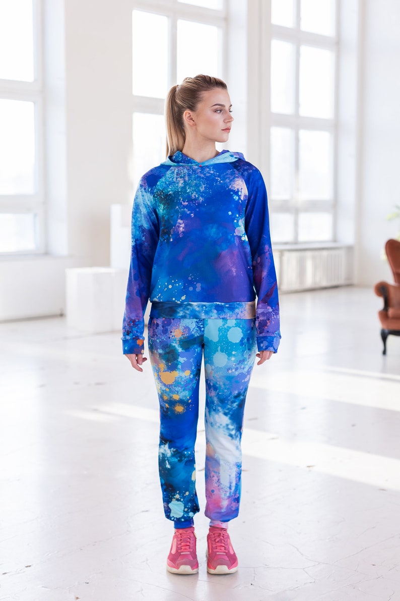 May include: A blue and purple hooded sweatshirt and matching pants with a paint splatter design. The model is wearing pink sneakers.