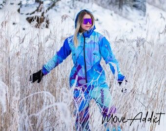 May include: A person wearing a blue and purple ski suit with a colorful abstract pattern. The suit has a hood and a zipper. The person is wearing black gloves and purple ski goggles. The person is standing in a snowy field with tall grass. The text "Move Addict" is visible in the lower right corner of the image.