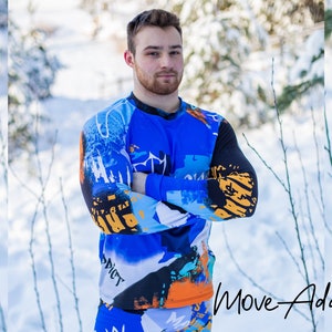 May include: A man wearing a blue, black and orange long-sleeved shirt with abstract designs and the word "addict" printed on it. The shirt has a crew neck and is made of a stretchy, comfortable fabric. The man is standing in a snowy woodland setting. The text "Move-Addict" is visible in the bottom right corner of the image.