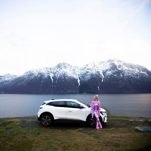 May include: A white SUV parked near a body of water with snow-capped mountains in the background. A person wearing a pink and purple patterned jumpsuit and boots is leaning against the car.