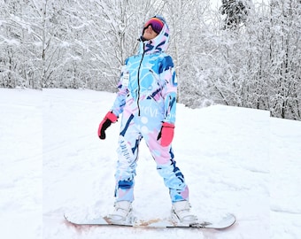 May include: A person wearing a blue and pink patterned ski suit with the words "Love Snow" on the leg, standing on a snowboard in a snowy forest.