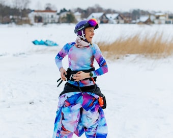 Op de afbeelding: Een vrouw in een kleurrijk skikostuum en helm staat op een besneeuwd veld. Ze past haar uitrusting aan en bereidt zich voor op wintersport.