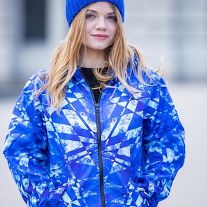 Könnte beinhalten: Eine blaue Softshelljacke mit geometrischem Muster in Weiß, mit Reißverschluss und Kapuze. Die Jacke wird von einer Person mit einer blauen Strickmütze getragen.