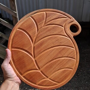 Puede incluir: Una tabla de servir redonda de madera con un diseño de hoja tallada. La tabla tiene un asa con un agujero.