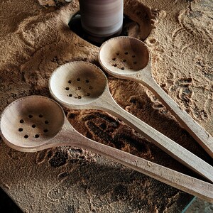 Puede incluir: Tres cucharas de madera con agujeros en el cuenco, que descansan sobre una superficie cubierta de aserr&iacute;n. Las cucharas est&aacute;n sin terminar y tienen un color de madera natural.
