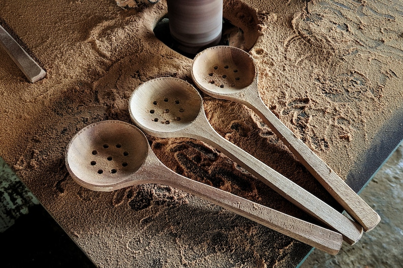 Puede incluir: Tres cucharas de madera con agujeros en el cuenco, que descansan sobre una superficie cubierta de aserr&iacute;n. Las cucharas est&aacute;n sin terminar y tienen un color de madera natural.