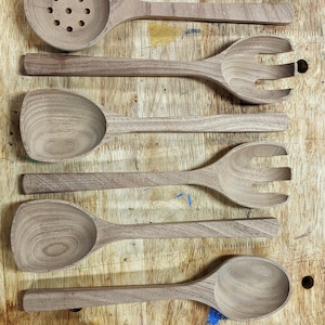 Puede incluir: Siete utensilios de cocina de madera, incluyendo cucharas, cucharones y una cuchara ranurada, est&aacute;n dispuestos en una fila sobre una superficie de madera. Los utensilios est&aacute;n hechos todos del mismo tipo de madera y tienen un acabado natural.