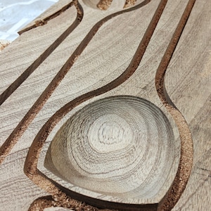 Puede incluir: Primer plano de una tabla de madera con dos formas de cuchara talladas. Las cucharas est&aacute;n sin terminar y tienen una veta de madera natural.