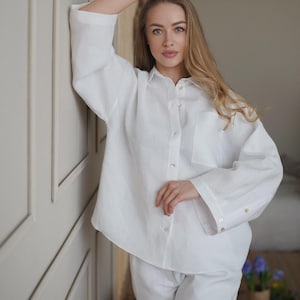 May include: A white linen shirt and matching trousers. The long-sleeved shirt has a button-down front and a chest pocket. The trousers appear to be loose-fitting. The outfit is made of a lightweight fabric.