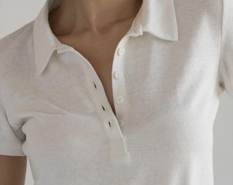 Pode incluir: Um close-up de uma camisa polo branca de mangas curtas. A camisa tem uma gola clássica e uma carcela de três botões. O tecido parece ser um material macio e texturizado. A camisa é de cor neutra, adequada para várias roupas.