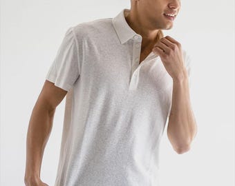 Pode incluir: Uma camisa polo cinza claro com mangas curtas e gola abotoada. A camisa tem uma aparência texturizada e é usada com calças cinza escuro. O modelo está tocando a gola da camisa.