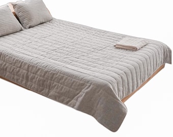 May include: A light grey quilted bedspread with matching pillowcases and a folded blanket. The bedspread and pillows have a textured, ribbed design. The bed is set on a wooden frame, creating a neutral and calming aesthetic.