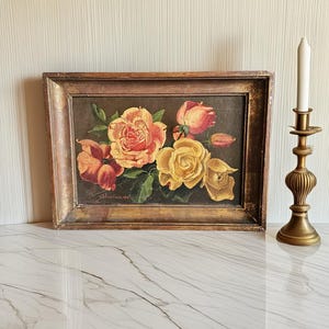 Könnte beinhalten: Ein rechteckiges Ölgemälde mit einem Strauß aus gelben, orangefarbenen und rosafarbenen Rosen mit grünen Blättern vor dunklem Hintergrund, in einem abgenutzten goldfarbenen Holzrahmen, begleitet von einem hohen Messingkerzenständer mit weißer Kerze.