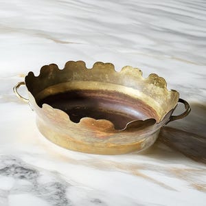 Pode incluir: Uma taça oval de latão com borda recortada e duas pequenas alças laterais. A superfície externa apresenta um acabamento dourado metálico desgastado com pátina visível, enquanto a base interna exibe um tom acobreado acastanhado mais escuro.