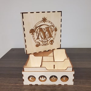 May include: A light-coloured wooden box with a lid, featuring laser-cut designs. The lid displays a circular emblem with the letter "M" and various symbols. The box is divided into compartments, and the front has circular designs. The box is made of wood.