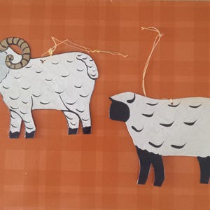 May include: Two flat, cutout sheep ornaments with gold hanging strings. The left sheep is white with brown horns and black hooves. The right sheep has a solid black head and black legs, with a white body featuring black curved markings.