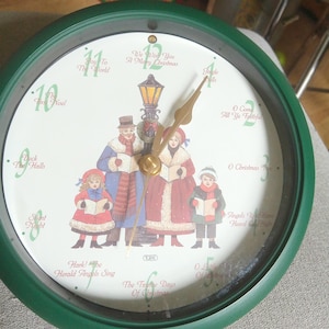 May include: A round wall clock with a dark green frame and a white face featuring a central illustration of a family caroling by a lamp post. The numbers are replaced by Christmas song titles including Silent Night and Jingle Bells.