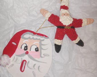 May include: Two Christmas ornaments: a Santa Claus face and a starfish-shaped Santa. The Santa face is painted on a white oval with a red hat and a fluffy white beard. The starfish Santa is red with white trim and black boots.