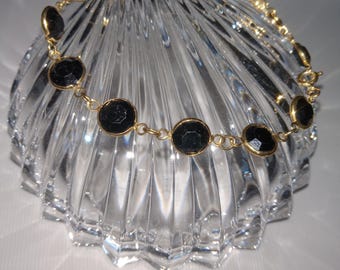 May include: A gold-toned chain bracelet featuring black, circular stones. The stones are set in gold bezels and connected by small gold links. The bracelet is displayed on a clear, textured glass dish.