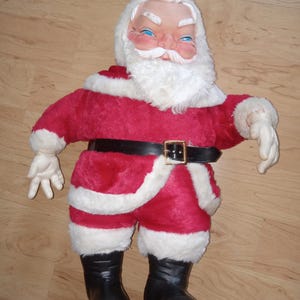 May include: A vintage Santa Claus doll with a red suit trimmed with white faux fur. The doll has a black belt with a gold buckle, black boots, and white gloves. The doll's face has blue eyes and a white beard.