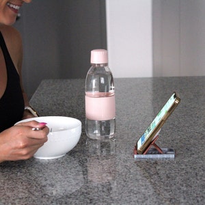 Op de afbeelding: Een doorzichtige waterfles met een roze deksel en een roze geribbelde gripsectie. Een witte kom en een smartphone in een standaard staan op een grijze aanrecht. De telefoon is naar de kijker gekanteld.
