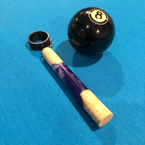 May include: A wooden pool cue stick with a purple and white swirl design. The stick has a black ring at the top and is resting on a blue felt pool table. A black eight ball is also on the table.