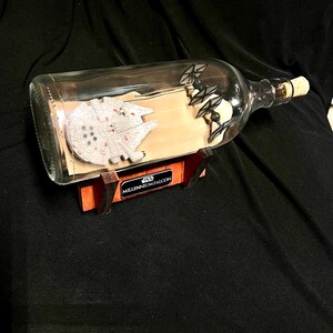 May include: A clear glass bottle with a cork stopper, containing a miniature Millennium Falcon model and TIE fighters. The bottle rests on a wooden stand with the words "Star Wars Millennium Falcon" printed on it. The background is a dark fabric.