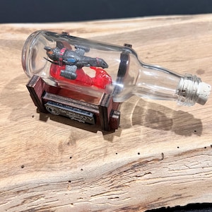 May include: A clear glass bottle with a cork stopper, containing a miniature spaceship model. The model is red and gray, and the bottle rests on a wooden stand with a metal plaque that reads "Serenity Firefly".