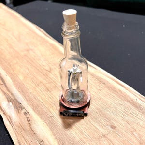 May include: A clear glass bottle with a cork stopper, containing a small white and gray figurine. The bottle is sitting on a wooden base with the text "VF-1S" printed on it.