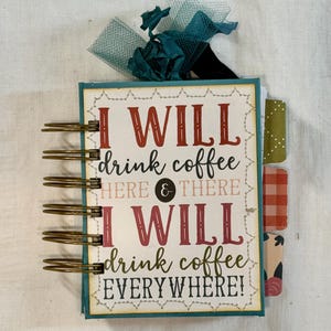 May include: A spiral-bound journal with the text "I WILL drink coffee HERE & THERE I WILL drink coffee EVERYWHERE!" in red and green lettering. The journal has decorative tabs and a teal ribbon.