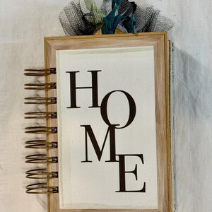 May include: A wooden framed notebook with the word "HOME" in large, dark letters on the front. The notebook is bound with a spiral coil and has a decorative ribbon on top. The frame is light brown and the background is white.
