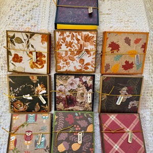 May include: A collection of ten square boxes with patterned lids, each wrapped in gold string and featuring a small metal charm with words including Faith, Hope, Love, Dream, Imagine, Believe, Inspire, and Enough, arranged in a grid pattern.