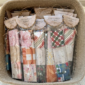 May include: A collection of rectangular paper packets in a woven basket, each featuring diverse patterns like floral, plaid, and musical notes. Every packet is sealed with a scalloped paper topper printed with the text A Little Something to Jot About.