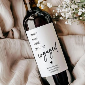 Może przedstawiać: Butelka wina z ciemnego szkła z białą prostokątną etykietą z czarnym napisem pairs well with getting engaged, małą ikoną serca oraz tekstem Congratulations Emma and Simon 06/08/2026, ozdobiona białymi kwiatami.