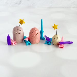 May include: Three small, rounded clay figures with smiling faces, two featuring yellow star toppers on wires. The figures are mauve, speckled pink, and speckled cream. They are surrounded by various small, colorful plastic tools in blue, purple, and pink.
