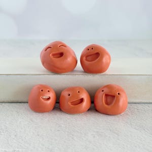 May include: Five small, round, terracotta-colored clay figurines, each featuring a unique carved facial expression. Two figures are positioned on a raised ledge, while three are arranged in a row below, all displaying various happy, smiling, or laughing faces.