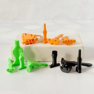 May include: An assortment of 3D-printed objects in various shapes and colors. The collection includes green, orange, and black pieces, some resembling abstract figures and others with geometric designs. The objects are arranged on a white surface.