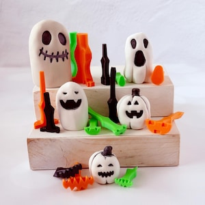 May include: A collection of Halloween-themed decorations, including white ghost figures with black facial features, orange pumpkins, and green and orange decorative elements. The decorations are arranged on a wooden display.