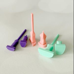 May include: A selection of brightly coloured, geometric objects in purple, pink, and mint green. The shapes include miniature tools and architectural forms. The objects are arranged on a light-coloured surface.