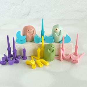 May include: A collection of small, colorful plastic tools and three rounded, smiling character figures in pink, light blue, and green. The set includes various pointed, pronged, and curved implements in purple, yellow, light blue, and pink shades.