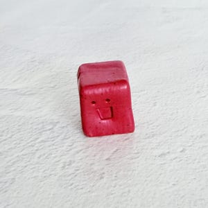 May include: A small, red, cube-shaped object with a smiling face carved into one side. The object has a slightly glossy finish and appears to be handmade. The background is a light, textured surface.