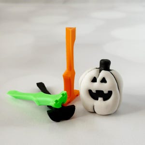 May include: A white pumpkin-shaped decoration with a black stem and carved face, next to an orange and green Halloween-themed decoration. The decorations are made of plastic and are on a white surface.