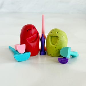 May include: A red rounded figure with a smiling face and a light green speckled rounded figure with a smiling face, accompanied by several small plastic tools in pink, purple, light blue, and teal colors arranged around the two figures.