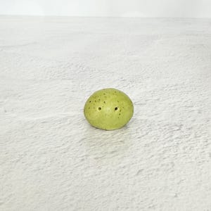 May include: A small, round, pale green ceramic salt shaker with two holes on top. The shaker has small, dark brown spots. The design is simple and minimalist, suitable for a kitchen or dining table.