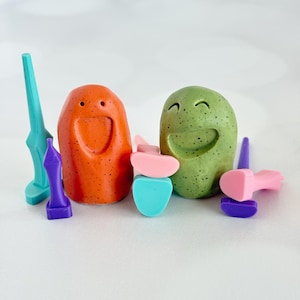May include: Two speckled clay figures, one orange and one green, each featuring a carved smiling face. They are surrounded by several small, colorful plastic tools in shades of teal, purple, and pink, scattered around the base of the figures.