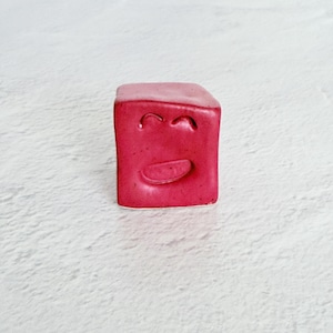 May include: A small, red cube with a smiling face carved into the front. The cube has rounded edges and a smooth surface. The background is a light, textured surface.
