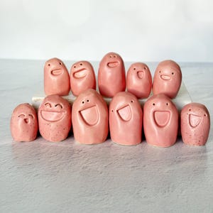 Puede incluir: Una colección de once figuras de cerámica rosa con caras sonrientes. Las figuras varían en tamaño y tienen diferentes expresiones, algunas con la boca abierta y otras cerradas. El fondo es una superficie gris claro.