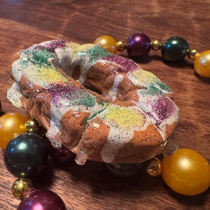 May include: A Mardi Gras-themed necklace featuring a king cake pendant and colorful beads. The king cake is decorated with white icing and sprinkles in green, yellow, and purple. The beads are in shades of gold, purple, and green.