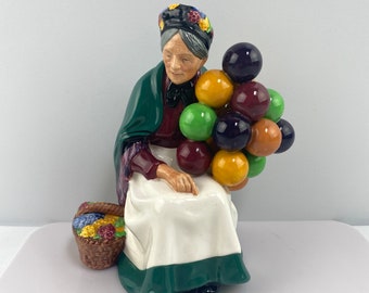 May include: A ceramic figurine of a woman wearing a green shawl and a floral hat, holding a bunch of colorful balloons. She is sitting down with a basket of fruit at her feet.